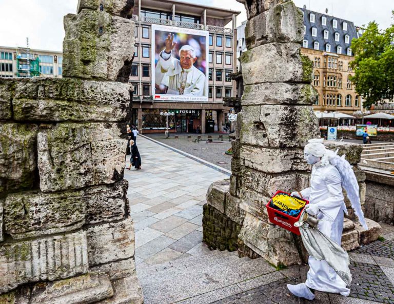 Ange avec caisse de bière devant le pape. Une statue vivante franchit la porte romaine devant la cathédrale de Cologne avec une caisse de bière qu'elle utilisera comme piédestal. En arrière-plan, le pape Benoît VI, plus grand que nature, fait signe depuis une affiche. / © Photo : Georg Berg